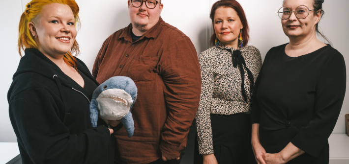 Vasemmaltta mustaan huppariin pukeutunut ja keltahiuksinen Elina hymyilee ja pitelee käsissään pientä hai-pehmolelua. Vieressä seisoo ruskeaan kauluspaitaan pukeutunut Kare, jonka vieressä pilkullisessa pailassa Sari, jolla on musta rusetti kaulassa ja korvissa sateenkaarikorvakorut. Oikealla laidalla seisoo kädet ristissä Maarit mustassa mekossa, hiukset nutturalla.