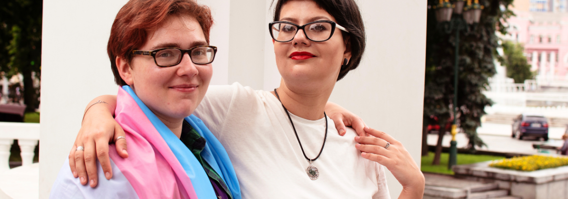 Nuori valkoihoinen pariskunta seisoo kädet toistensa olkapäillä ulkona vakoisen rakennelman edessä. Molemmilla on silmälasit ja lyhyet hiukset. Toisella on harteillaan translippu.