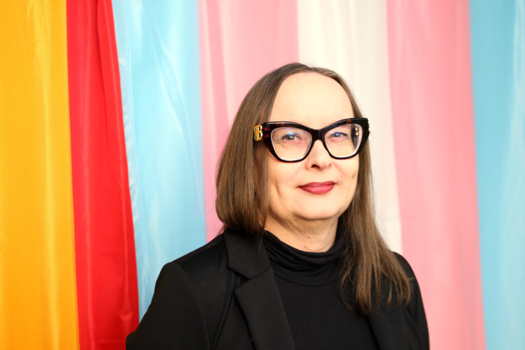 Translipun ja sateenkaarilipun edessä seisova Maarit Huuska. Hänellä on pitkät ruskeat hiukset, musta jakku ja poolopaita sekä mustat näyttävät silmälasit.